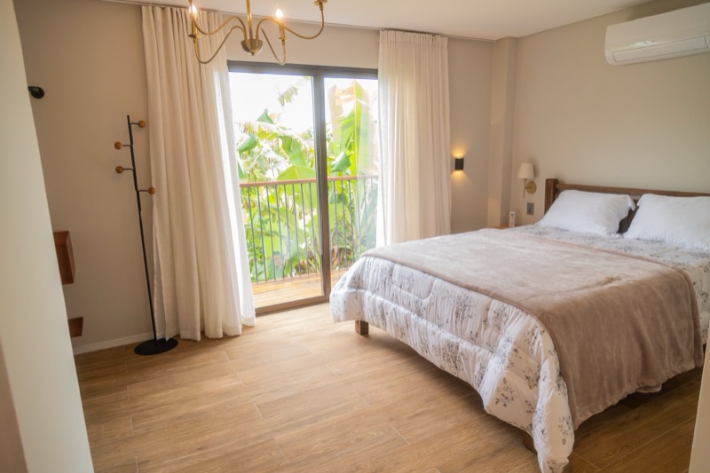 Cama queen com colcha estampada de flores e a porta de vidro aberta para o deck da varanda, com as bananeiras lá fora