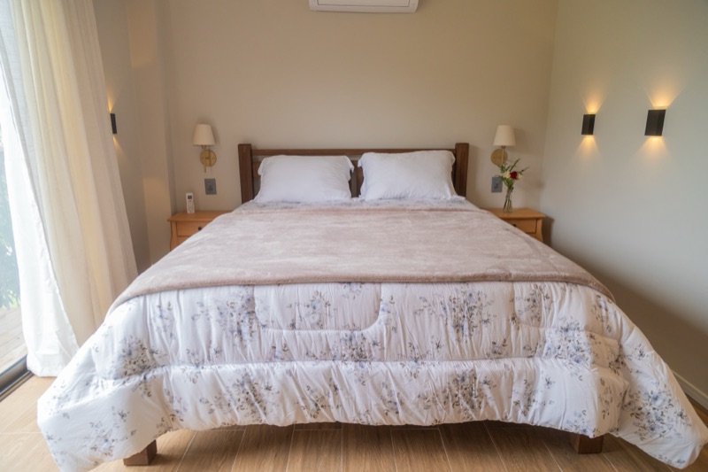 Cama queen de frente, com cabeceira de madeira, arandelas acesas dos dois lados e uma flor em cada mesa de cabeceira