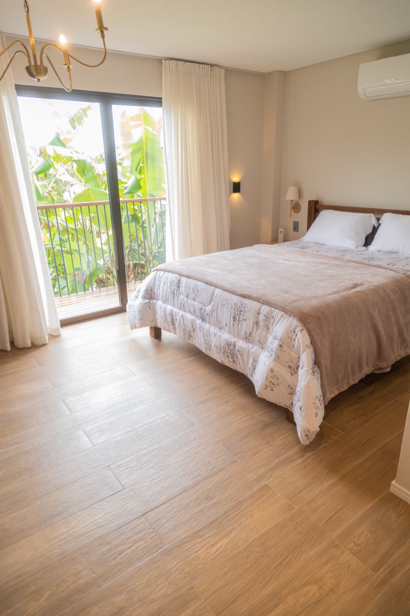 Cama queen à direita e, à esquerda, a porta de vidro da varanda aberta para o verde, sobre piso de madeira