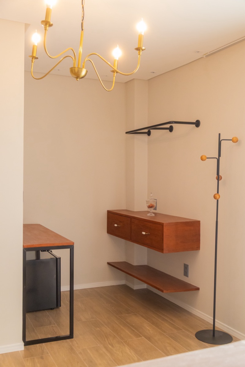 Canto do closet, com cabideiro de parede, gaveteiro suspenso de madeira, cabideiro de pé e a escrivaninha com o frigobar embaixo