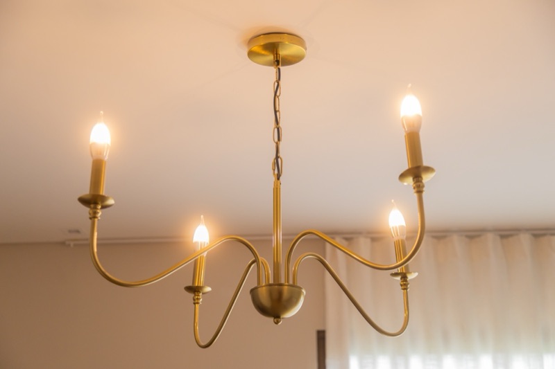 Lustre dourado de cinco braços, com lâmpadas em formato de vela, e a cortina ao fundo