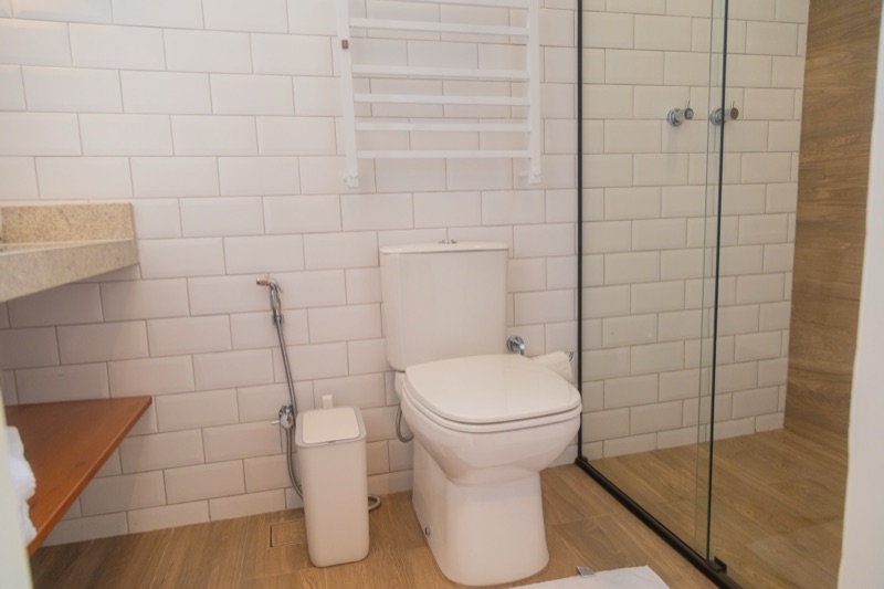 Banheiro com azulejo branco, aquecedor de toalhas na parede e box de vidro com parede de porcelanato amadeirado